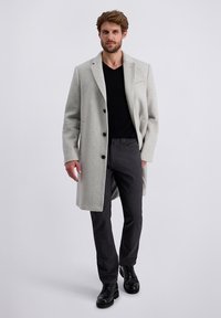 Lichtgrijze wollen overjas met zwarte knopen, geknipte revers en zijzakken, gecombineerd met een zwarte v-halstrui en donkergrijze broeken. Zwarte laarzen.