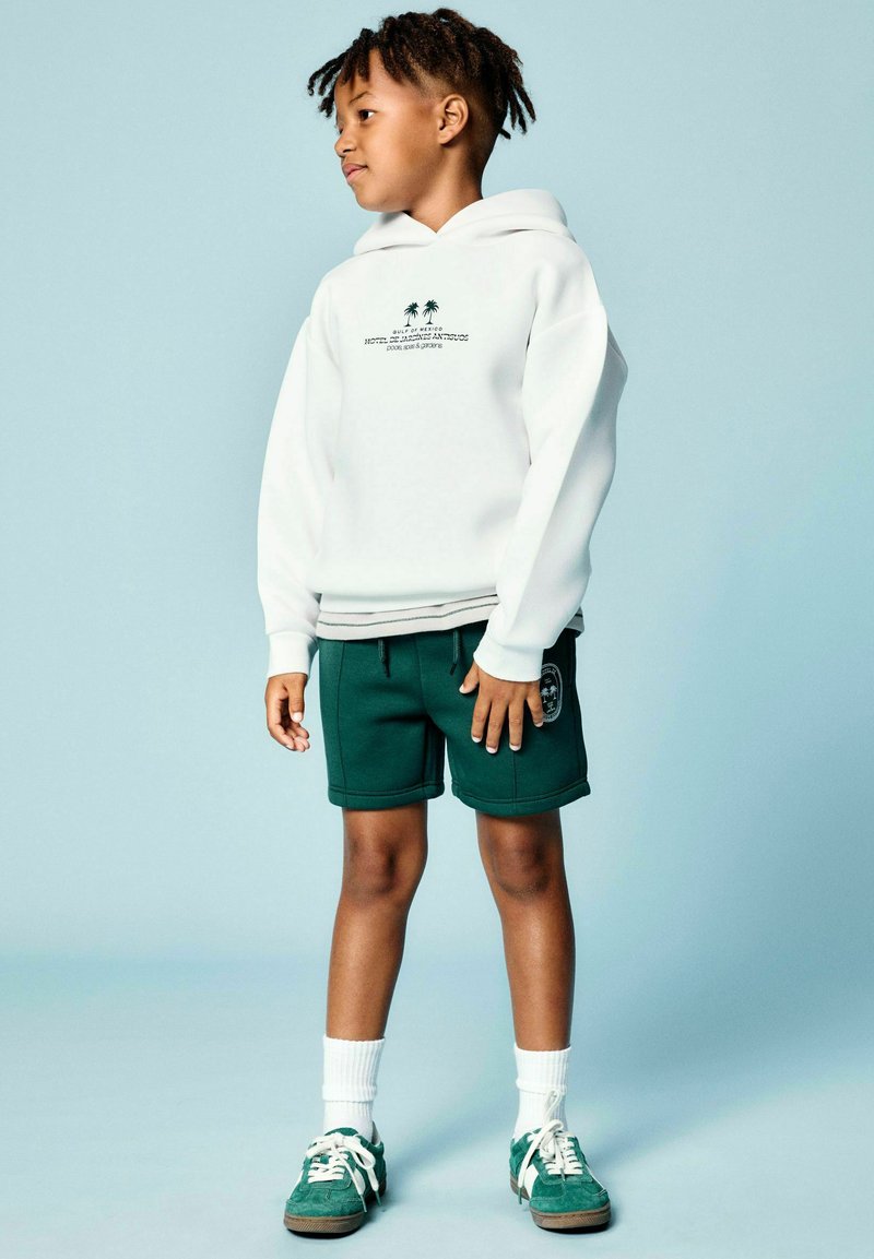 Garçon debout portant un sweat à capuche blanc avec un motif de palmier, un short vert foncé, des chaussettes blanches et des baskets vertes sur un fond bleu clair.