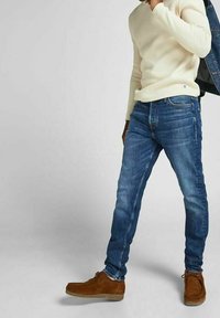 Tmavě modré džínové kalhoty, slim fit, s jemným zesvětlením, v kombinaci s krémovým texturovaným svetrem a hnědými semišovými botami.