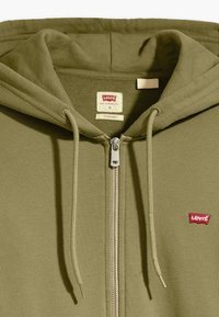 Olivengrønn hettejakke med glidelås laget av teksturert stoff, med snor og liten rød Levi's-logo på venstre bryst og hette.