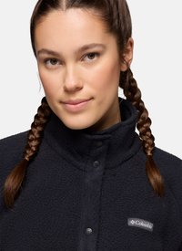 Jonge vrouw met bruin gevlochten haar die een zwarte, gestructureerde Columbia fleecejas met een hoge, met knopen gesloten kraag draagt, kijkt vooruit.