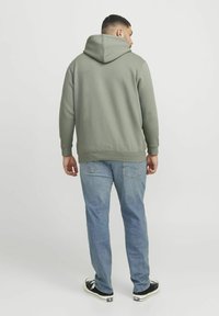 Grøn hoodie med stor hætte på ryggen, ribbede ærmer og forneden, parret med lyseblå jeans og sorte sneakers. Ensfarvet baggrund.