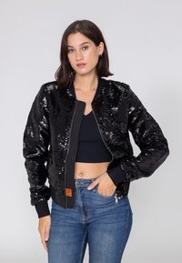 Jonge vrouw met een zwart pailletten bomberjack, een zwart cropped top en een blauwe spijkerbroek, poseert met één hand in de zak tegen een effen achtergrond.