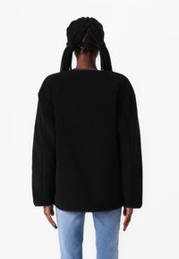 Chaqueta de forro polar negra con un corte holgado, hombros caídos y acabado texturizado. Combinada con vaqueros azul claro, vista desde atrás.