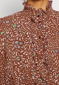 Braun gemusterte Bluse mit einem Rüschenausschnitt, weißen Blumenmotiven und braunen Knöpfen entlang der Knopfleiste, aus leichtem Stoff gefertigt.