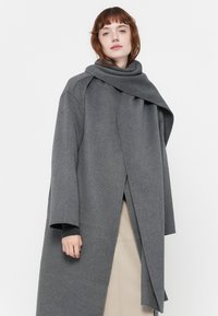 Grijze wollen jas met lange mouwen, oversized kraag en een gedrapeerd design. Voorzien van een voorste wikkeldetail en zachte textuur.