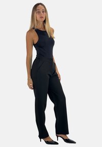 Zwarte op maat gemaakte broek met een hoge taille, plooien en een knoopsluiting. Gecombineerd met een aansluitend zwart hemdje en zwarte puntige hakken.