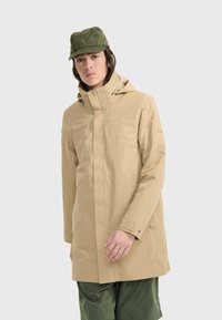 Giacca impermeabile beige con cappuccio, dotata di chiusura frontale a zip e polsini con bottoni. Abbinata a pantaloni cargo verde oliva e un cappello coordinato.