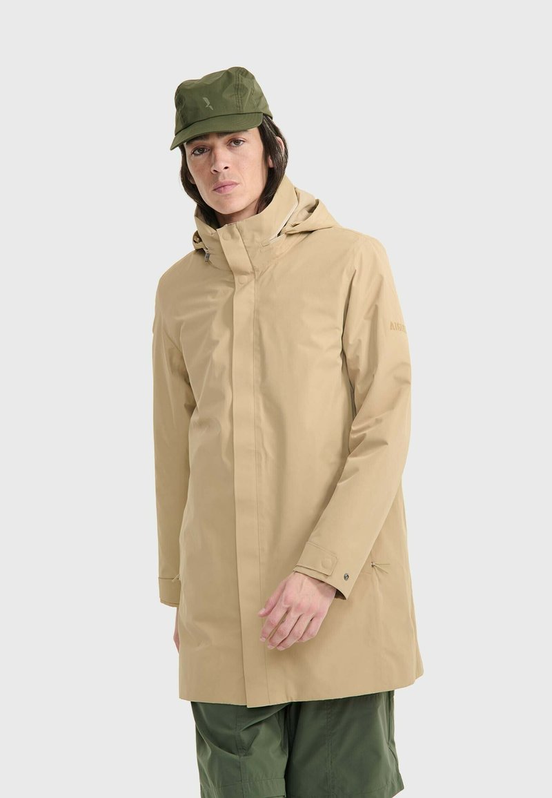 Giacca impermeabile beige con cappuccio, dotata di chiusura frontale a zip e polsini con bottoni. Abbinata a pantaloni cargo verde oliva e un cappello coordinato.