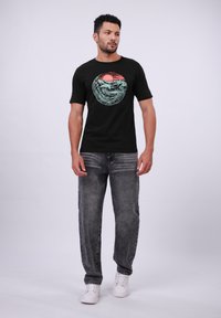 Zwarte t-shirt met een kleurrijke golf- en zonafbeelding, gecombineerd met vervaagde grijze spijkerbroek en witte sneakers. De outfit heeft korte mouwen en een relaxte pasvorm.