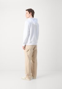 Felpa bianca con cappuccio regolabile, abbinata a pantaloni beige larghi. Sneakers bianche con dettagli beige. Estetica streetwear semplice e casual.