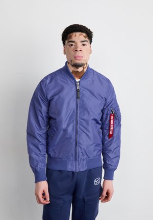 Giacca bomber blu con una texture leggera e lucida, chiusura con zip, polsini a costine e una patch ben visibile sulla manica sinistra. Abbinata a pantaloni blu navy.
