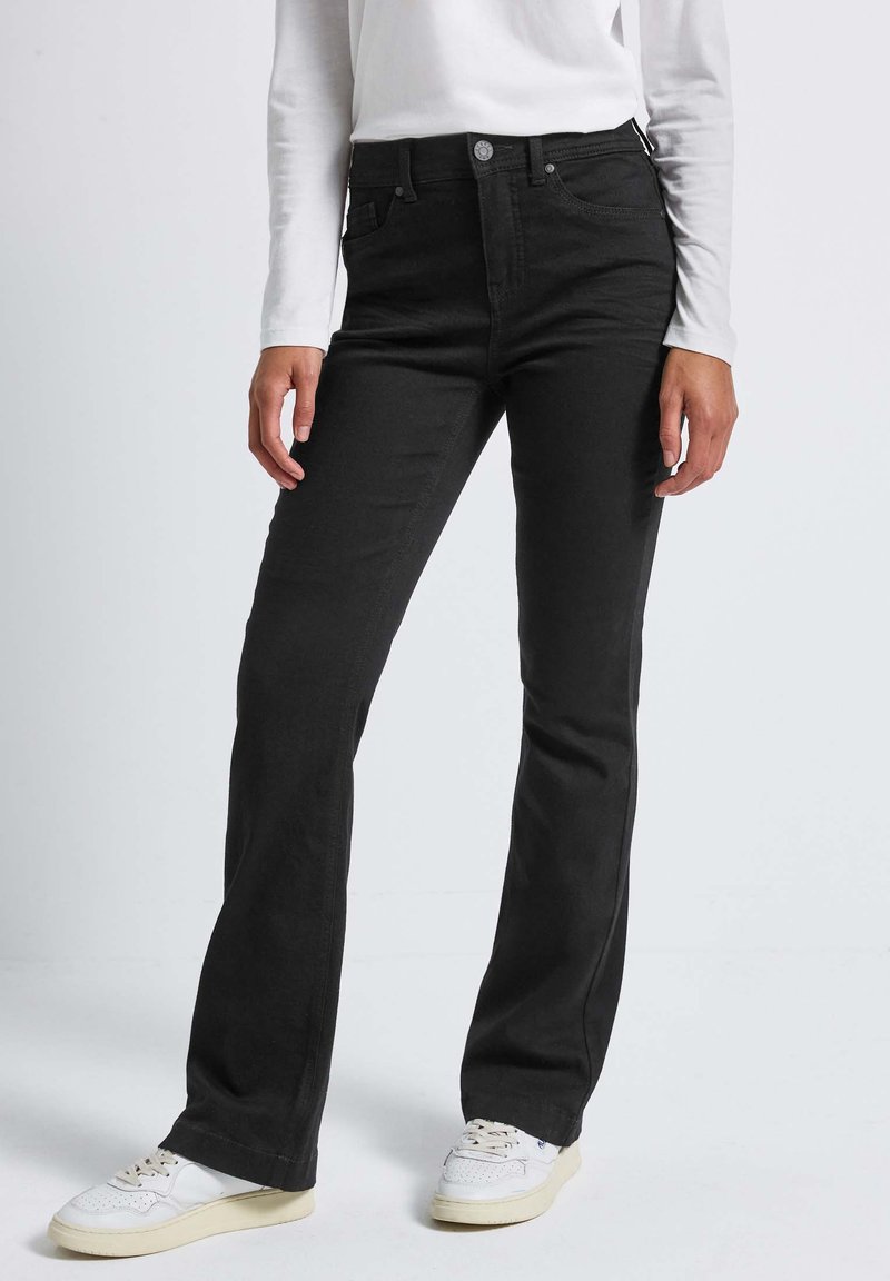 Jeans en denim noir évasés avec une taille haute, dotés de poches à l'avant et d'un zip. Portés avec des baskets blanches, sur un fond clair.