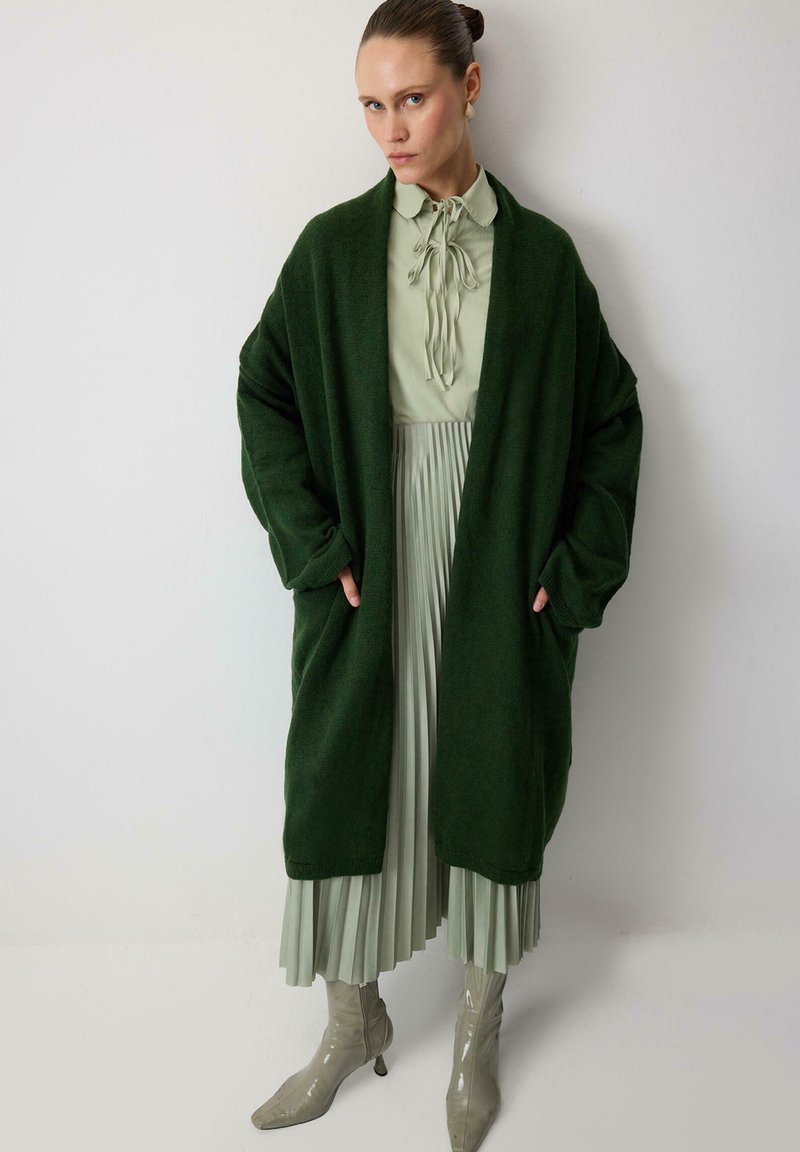 Dark green oversized cardigan in soft fabric, layered over a light green pleated dress with a tie-neck, paired with glossy beige ankle boots.