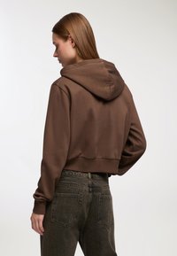 Donna con capelli castani lisci volta di lato, indossa un cropped hoodie marrone e jeans neri sbiaditi a vita alta contro uno sfondo semplice.