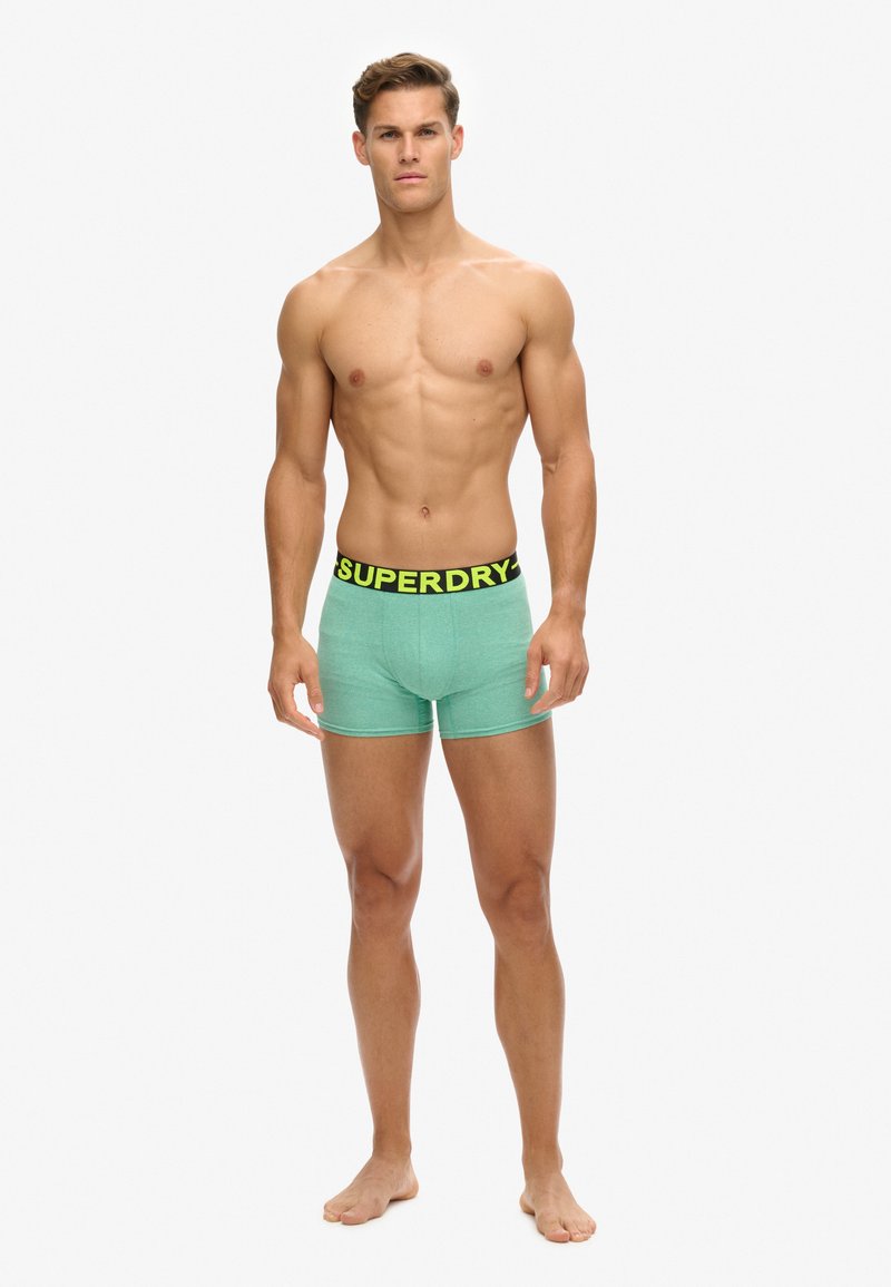 Bóxers verdes con una cinturilla de logo negra que presenta "SUPERDRY" en un amarillo brillante. La tela parece suave y tiene un diseño ajustado.