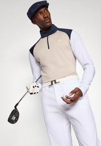 Μπεζ και ναυτικό μπλε μακριμάνικο polo γκολφ με κολάρο με φερμουάρ, λευκά παντελόνια, λευκή ζώνη με λογότυπο, κρατώντας μια γκολφομπαλά και μπαστούνι γκολφ, με γάντια με υφή.