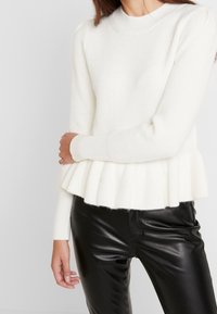 Valkoinen ribbineulottu neule pyöreällä pääntiellä, pitkät hihat ja peplum-helma. Yhdistetty mustiin nahkahousuihin, joissa on teksturoidu pinta.