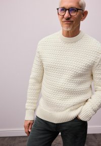 Weißer gestrickter Pullover mit einem strukturierten, dicken Muster sowie gerippten Bündchen und Saum. Kombiniert mit dunkelgrauen Hosen.