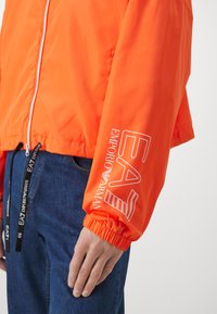 Lys orange zip-jakke med elastiske manchetter, med hvidt trykt logo på ærmet og snoretræk i kanten. Sammen med blå denim.