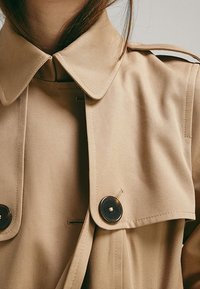 Beige trench coat featuring a classic collar, double-breasted design, and large black buttons. Made from smooth, lightweight fabric.