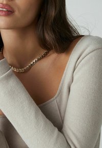 Šviesiai pilkas banguotas ilgomis rankovėmis megztinis su plačiu iškirpimu. Auksinė suktinė grandinė priglunda prie odos, suteikdama metalinį akcentą.