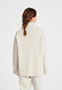 Licht beige oversized trui met een hoge geribbelde kraag en geribbelde manchetten. Zachte textuur en ontspannen silhouet. De achterkant toont een gevlochten haarstijl.