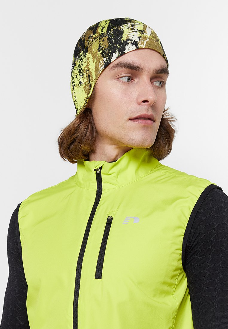 Zielono-czarna opaska na głowę noszona z neonowożółtą kamizelką. Kamizelka ma kieszeń na zamek oraz elegancki, dopasowany krój.