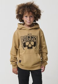 Dieťa s kučeravými vlasmi, oblečené v béžovej mikine s kapucňou, na ktorej je lebka a nápis "RUGGED", stojí na svetlom pozadí.