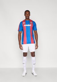 Κόκκινη και μπλε κάθετη ριγέ φανέλα ποδοσφαίρου με λευκό σορτς, κάλτσες Nike και παπούτσια ποδοσφαίρου· διαθέτει το λογότυπο MaxBet και τη σήμανση Kappa.