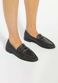 Black leather loafers with silver studs, fringe detail, flat soles, and slip-on design. Smooth texture and rounded toe shape.