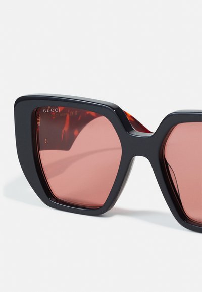 Gucci zonnebril met gedurfde zwarte monturen in hoekige vormen, rozenkleurige lenzen en een glanzende afwerking met logo-detail aan de zijkant.