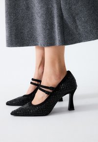 Zapatos negros de tacón alto con punta afilada, diseñados con un material sintético texturizado y dobles correas en el tobillo. Detallados con tachuelas elevadas.