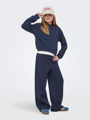 Meitene tumši zilā sporta krekla un platām biksēm, pieskaras bēšai cepurei ar sarkanu logotipu, valkā zilas kurpes, stāv uz gaiša fona.