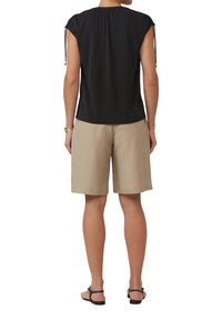 Schwarzes kurzärmliges Oberteil mit gerafftem Rücken, kombiniert mit beigen Leinen-Shorts. Das Outfit hat verstellbare Bindebänder an den Ärmeln und flache Sandalen.