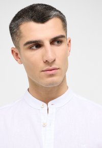 Camicia bianca di lino con colletto alla cinese, dotata di una plica con due bottoni; texture fine, dettagli di cucitura discreti e senza pattern visibili.