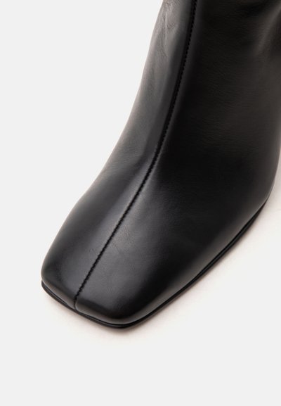 Gros plan d'une botte en cuir noir avec un bout carré et une couture centrale visible sur fond blanc.