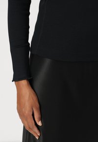 Black long-sleeve top with a fitted design and raw edge cuffs, paired with a smooth black textured skirt. Showing part of a hand.