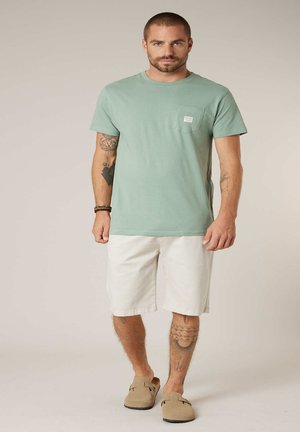 Man met tatoeages die een groene T-shirt met korte mouwen, witte knielange shorts, beige instapschoenen en armbanden draagt tegen een neutrale achtergrond.