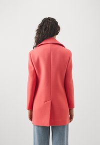Abrigo de lana color coral con cuello de muesca, silueta recta y una abertura trasera, combinado con unos vaqueros de denim azul claro.