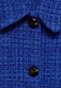 Modrý textil s tkaným vzorem, na němž je černý knoflík a límec. Detailní záběr ukazuje bohatou strukturu materiálu.