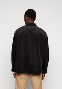 Fekete kordbársony ing, laza szabással, gallérral, hosszú ujjaival és gombos mandzsettával. Texturált anyag, függőleges bordákkal.