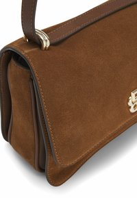 Brun ruskindshåndtaske med en guldfarvet metaldetalje, der har en klaplukning og en brun læderrem, som fremviser en blød tekstur og en struktureret form.