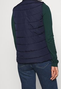 Persoon met een marineblauw gestept vest over een groene lange mouwen shirt en blauwe spijkerbroek, gezien van achteren, staand tegen een effen achtergrond.