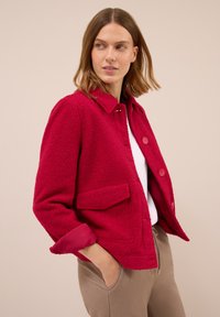 Femme portant une veste texturée rouge vif sur un haut blanc et un pantalon beige, regardant sur le côté avec les mains dans les poches devant un fond neutre.