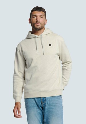 Lichte beige hoodie van zacht fabric, met een trekkoordkap en een hexagonaal logo accent. Gekleed met lichtblauwe spijkerbroek.