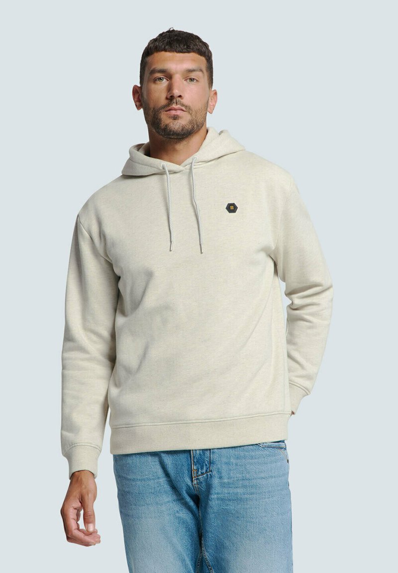 Lichte beige hoodie van zacht fabric, met een trekkoordkap en een hexagonaal logo accent. Gekleed met lichtblauwe spijkerbroek.