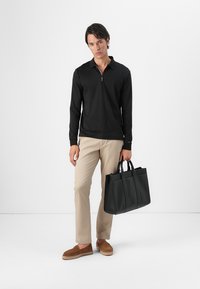 Camisa polo negra de manga larga con cuello con cremallera, pantalones beiges, espadrilles en color marrón, sosteniendo un bolso de mano de cuero negro con doble asa.