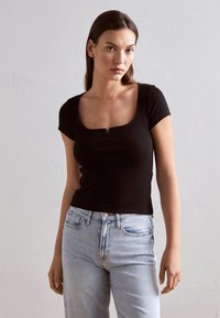 Top corto a maniche corte nero a coste con scollatura quadrata e un piccolo dettaglio a taglio, abbinato a jeans in denim blu chiaro.
