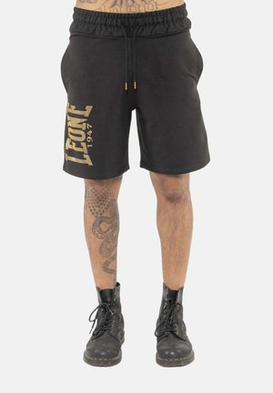 Short noir avec texte jaune "Leone 1947" porté par une personne tatouée en bottes noires de combat, debout sur fond blanc.
